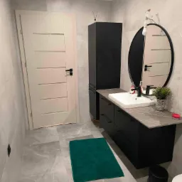 Nowoczesna łazienka z czarnymi meblami, owalnym lustrem i szarą płytką. Uwagę przyciąga intensywnie zielony dywanik, dodający kontrastu kolorystycznego wnętrzu.