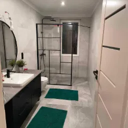 Nowoczesna łazienka z szarymi płytkami, kabiną prysznicową typu walk-in z czarnymi profilami, czarną szafką pod umywalką i zielonymi dywanikami.