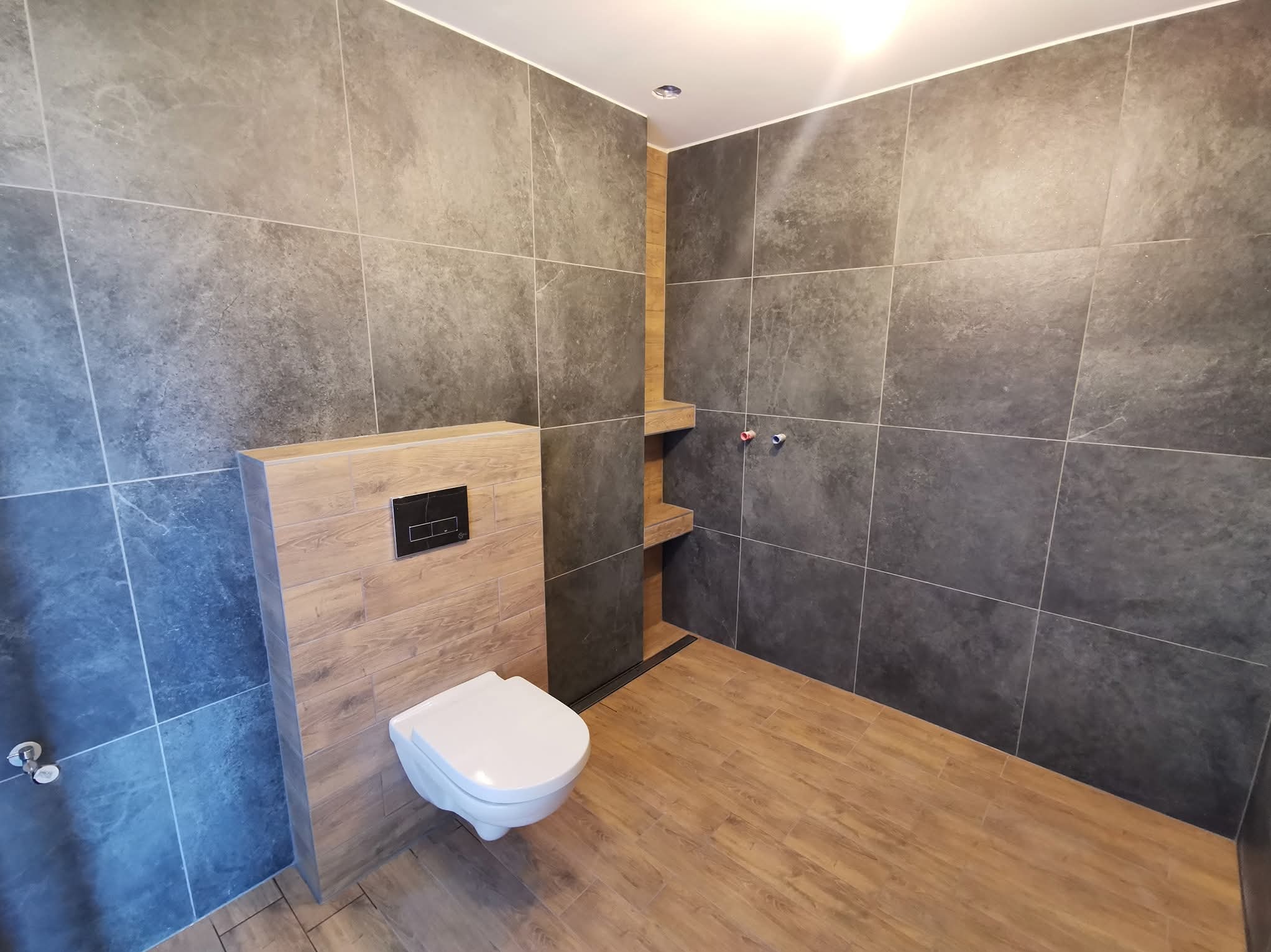 Wykończona łazienka z szarymi płytkami ściennymi, drewnopodobną podłogą i toaletą podwieszaną. Widoczne półki wnękowe i minimalistyczny design.