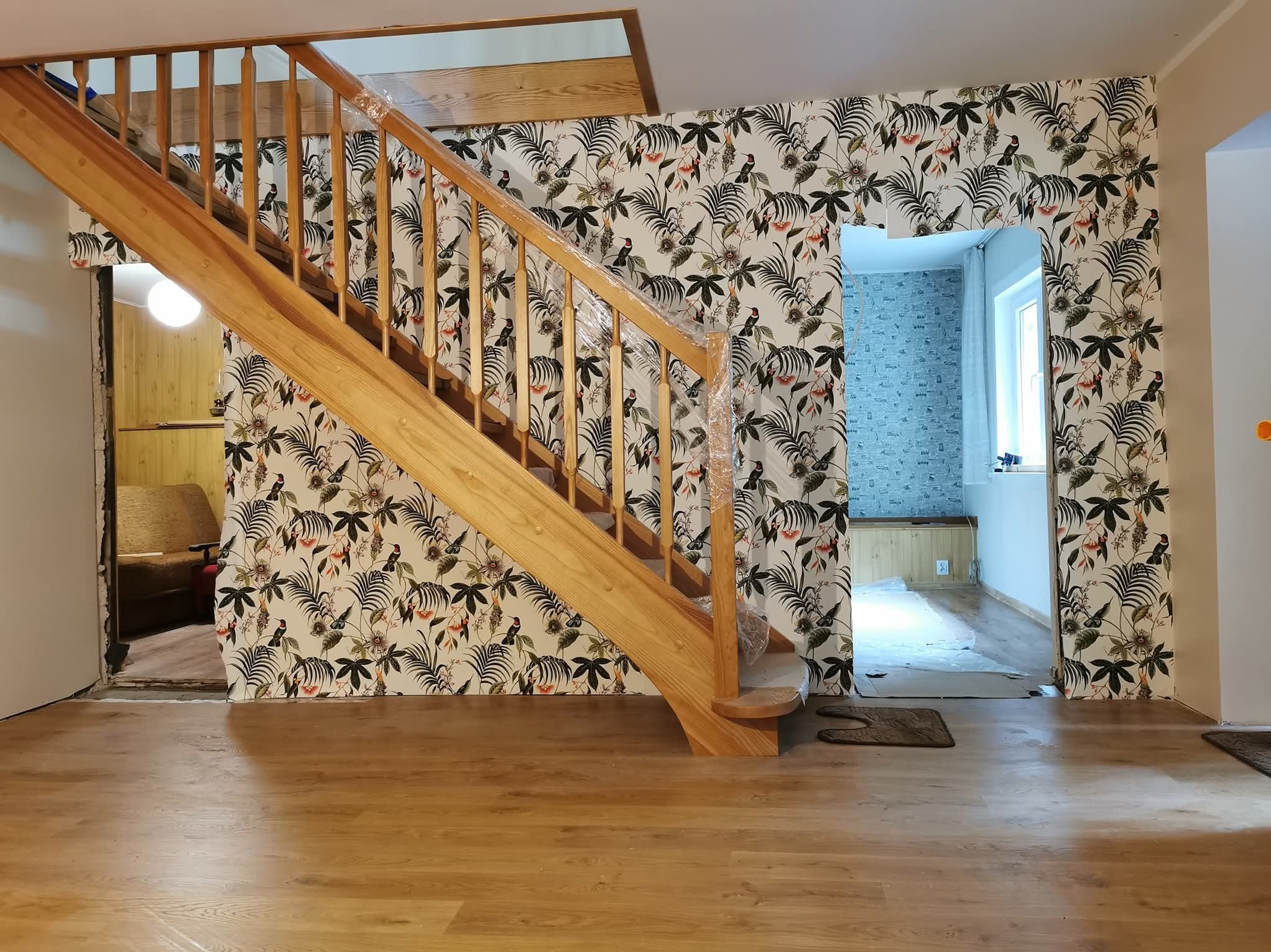 Jasne drewniane schody z zabezpieczeniem folią, wzorzysta tapeta w tropikalne liście i ptaki na ścianie, panele podłogowe w odcieniach brązu. Widoczne przejścia do innych pomieszczeń.