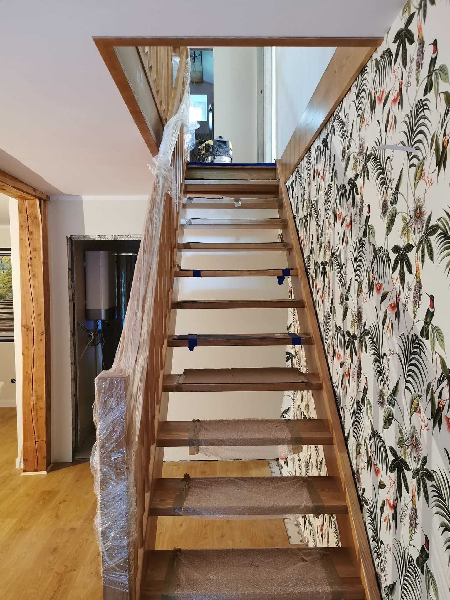 Drewniane schody w trakcie montażu, zabezpieczone folią i taśmą. Ściana z tapetą w tropikalne wzory. Widoczne elementy drewniane i instalacja w tle.