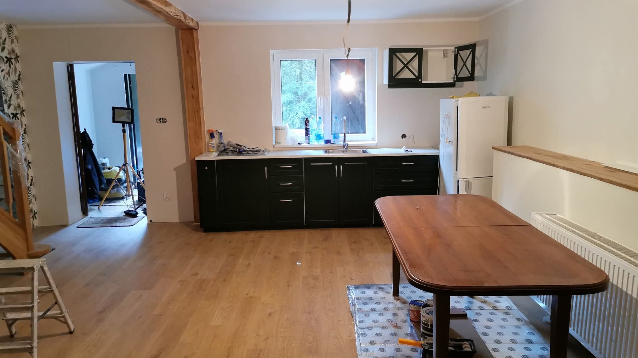 Jasne wnętrze kuchni po remoncie: drewniana podłoga, ciemne szafki kuchenne, biała lodówka i okno z widokiem na zieleń. Częściowo widoczny stół i drabina.