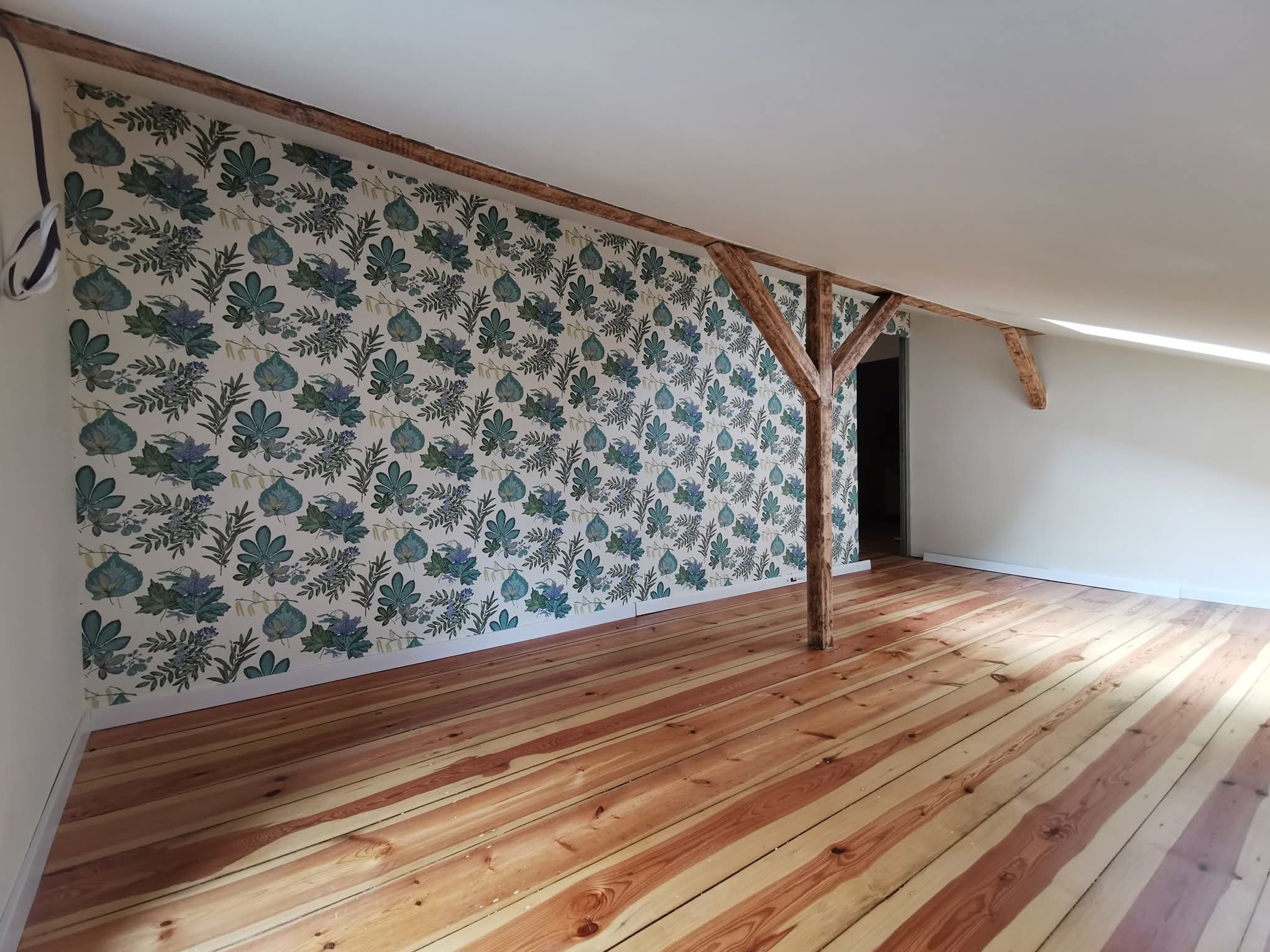 Pokój na poddaszu po remoncie: drewniana podłoga, ściana z wzorzystą tapetą, drewniane belki konstrukcyjne, sufit skośny. Widok na odnowione wnętrze.