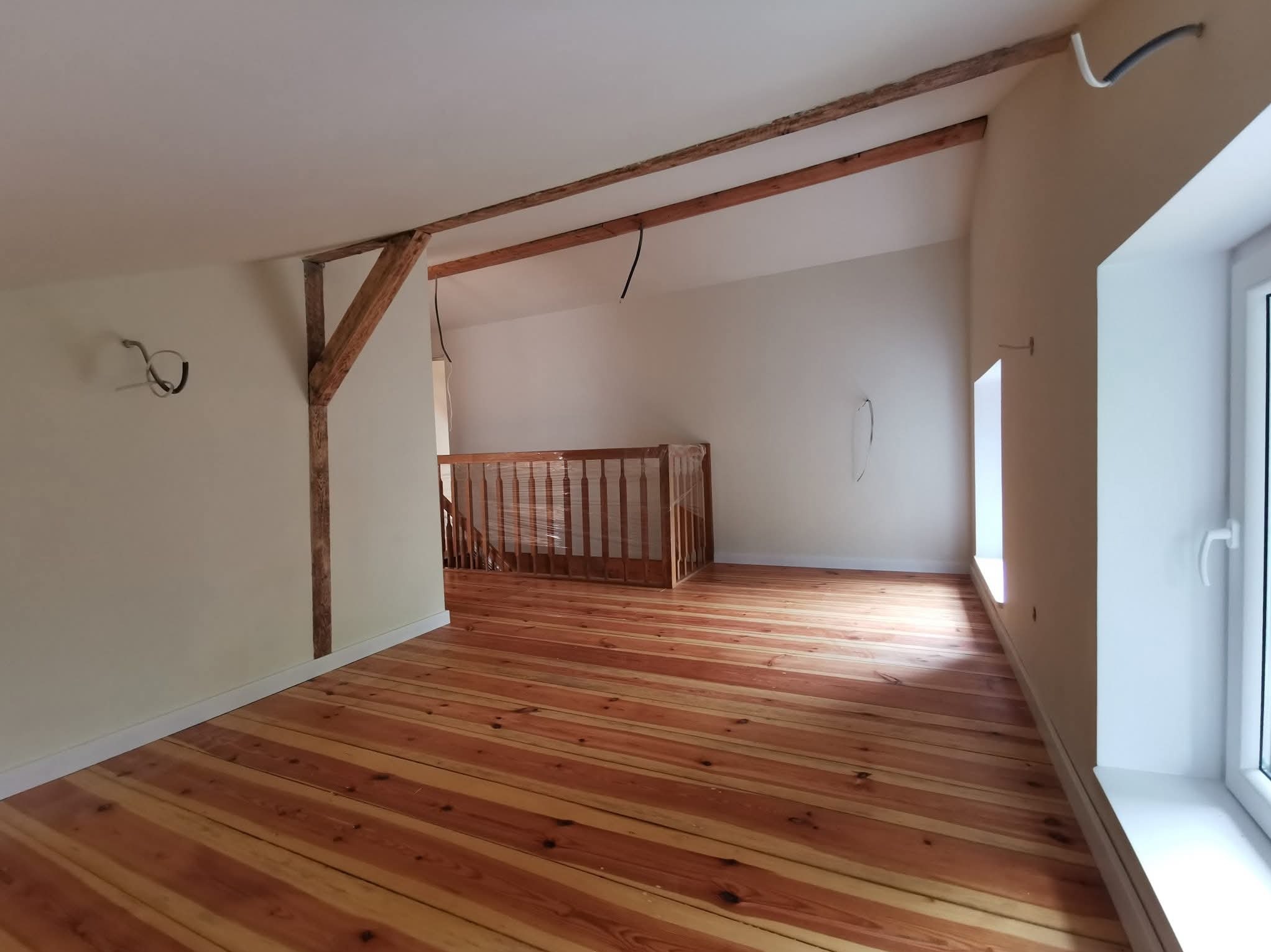 Jasne, przestronne poddasze po remoncie: drewniana podłoga, pomalowane ściany, widoczne belki stropowe i balustrada schodów na antresolę. Częściowo widoczne kable.