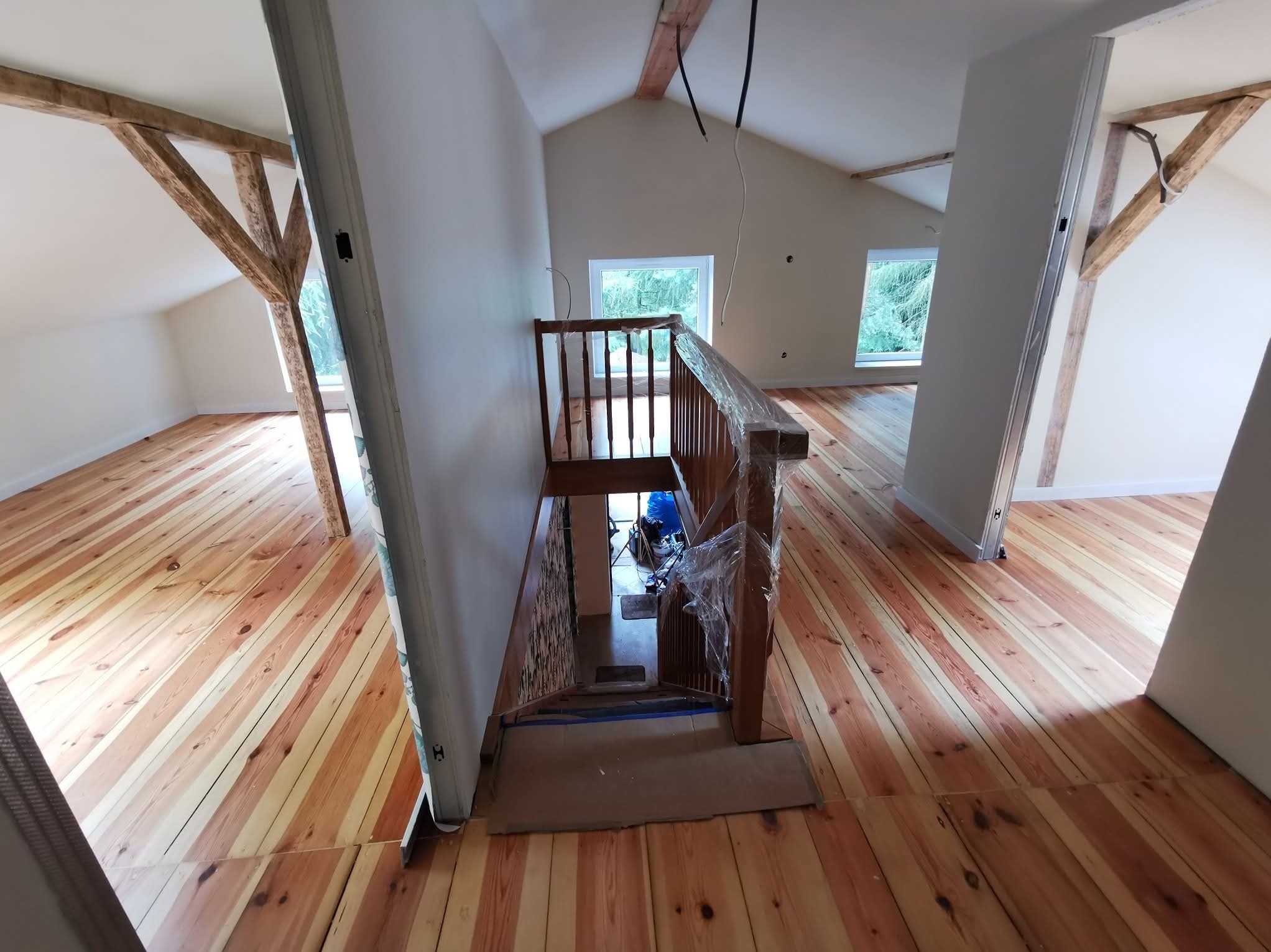 Poddasze po renowacji: świeżo polakierowana drewniana podłoga ułożona pod skosem, białe ściany i drewniane belki, schody zabezpieczone folią. Widok z góry na dół.