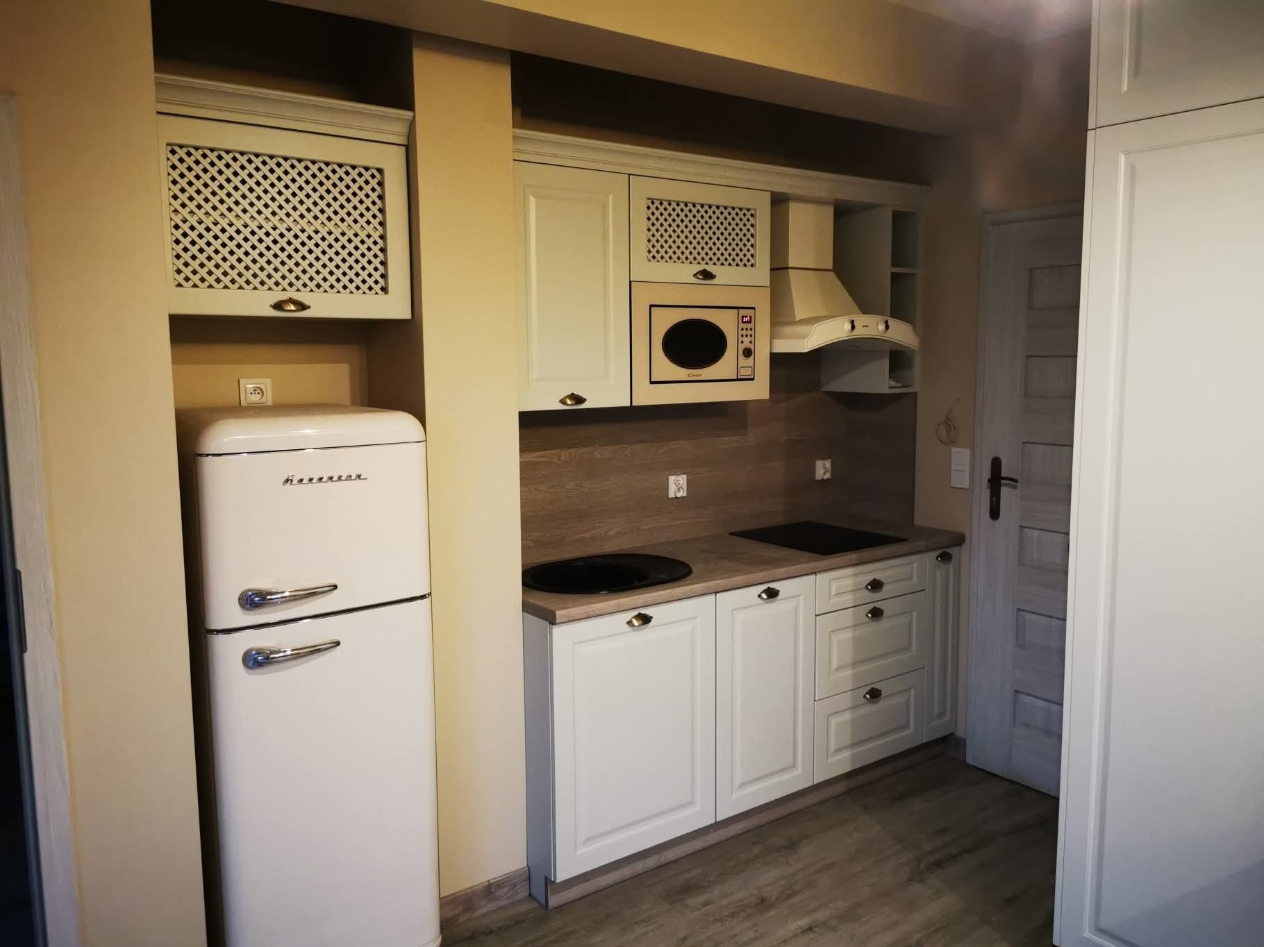 Jasna kuchnia z kremowymi szafkami, retro lodówką i drewnianym blatem. Górne szafki zdobione kratką. Ujęcie prezentuje gotową aranżację.