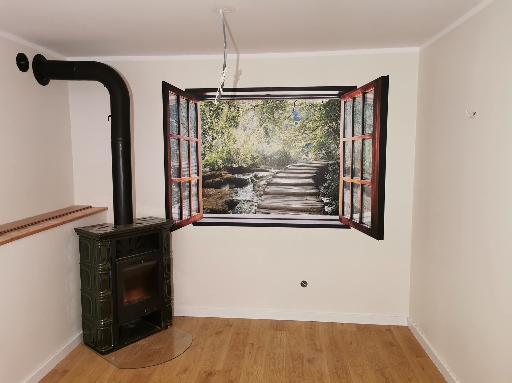 Pokój z piecem kaflowym i fototapetą imitującą okno z widokiem na las i wodospad, parkiet na podłodze, jasne ściany.