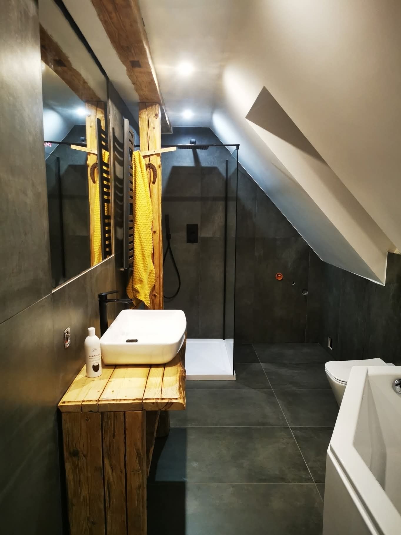 Nowoczesna łazienka na poddaszu z kabiną prysznicową, umywalką na drewnianym blacie i szarą glazurą. Widoczne drewniane belki stropowe i żółty ręcznik.