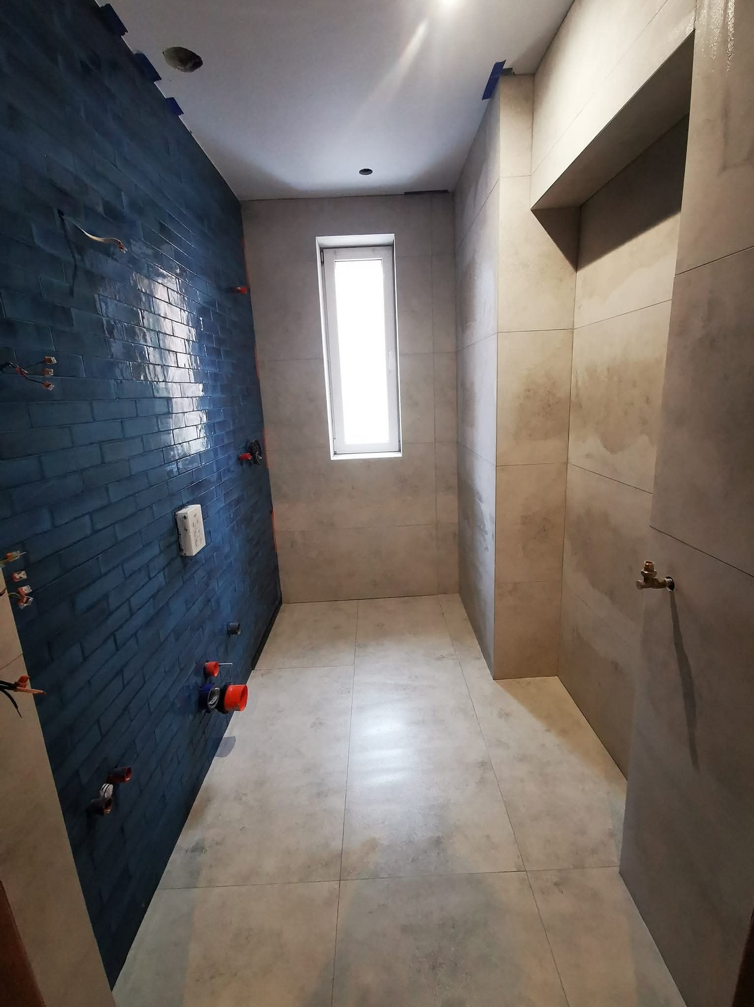 Nowoczesna łazienka w trakcie wykańczania: ściana z ciemnych, błyszczących płytek, jasne płytki na pozostałych ścianach i podłodze, widoczne instalacje wodne.