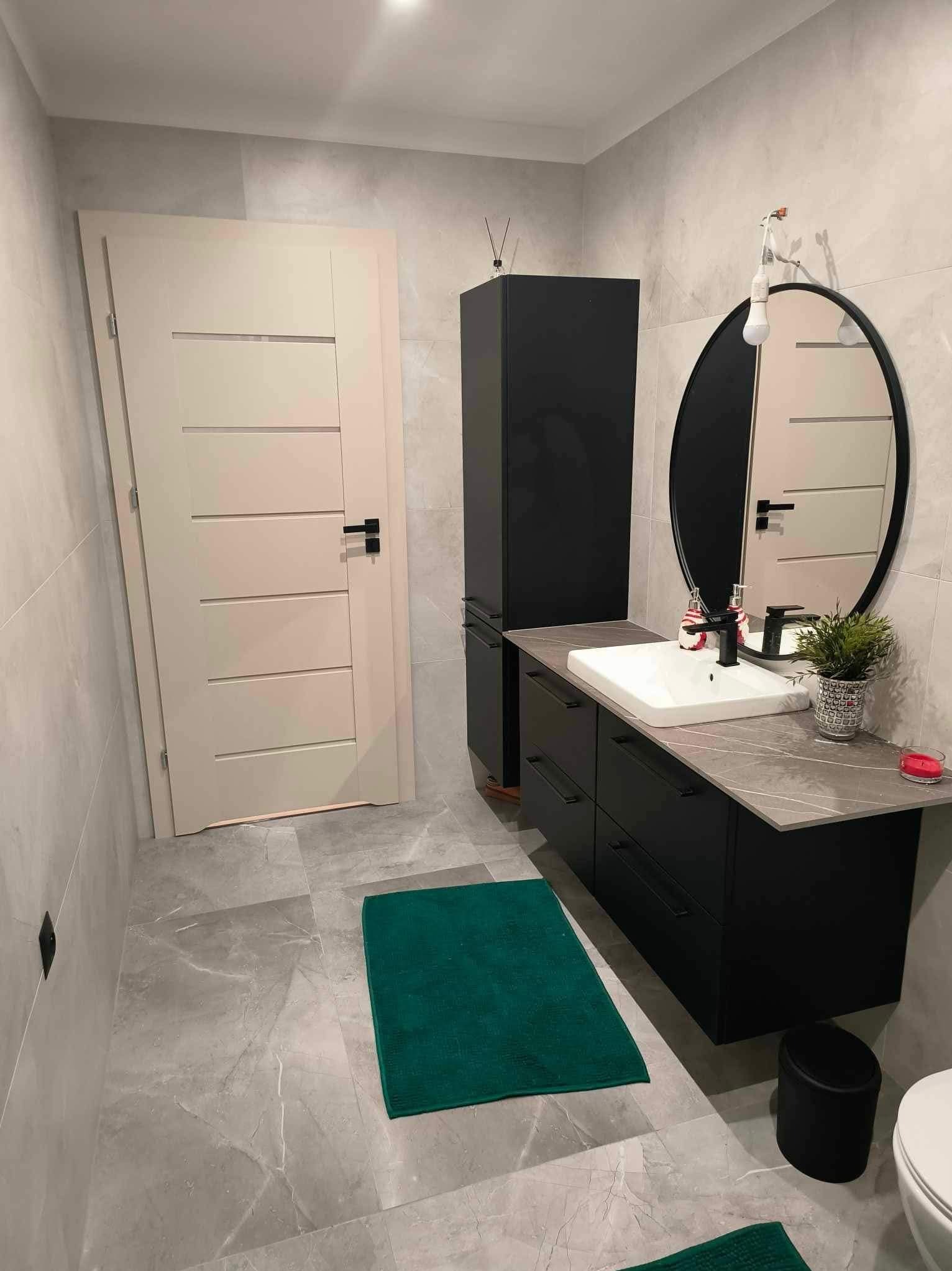 Nowoczesna łazienka z czarnymi meblami, owalnym lustrem i szarą płytką. Uwagę przyciąga intensywnie zielony dywanik, dodający kontrastu kolorystycznego wnętrzu.