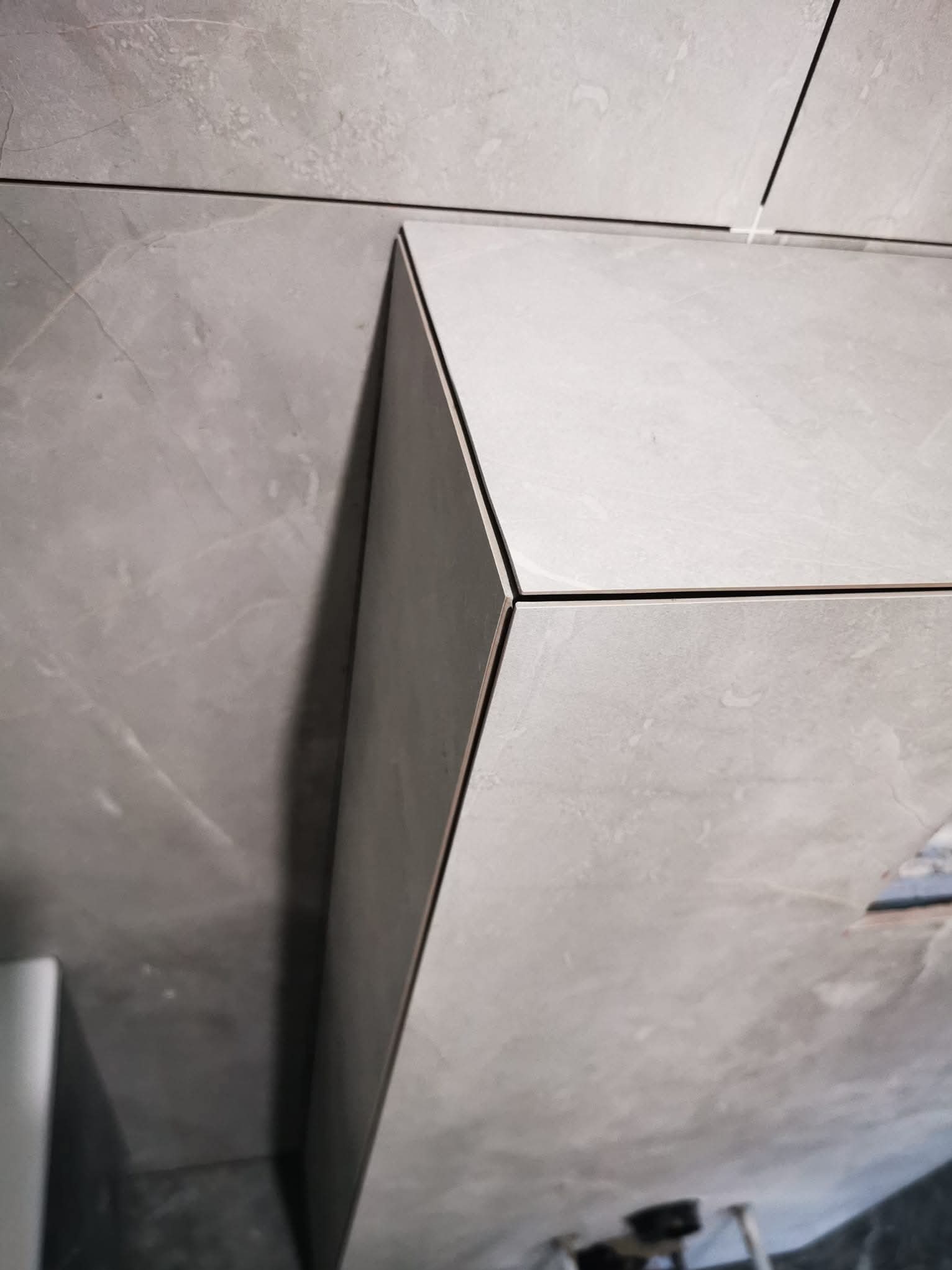 Szare, matowe płytki imitujące marmur, ułożone z precyzją na ścianie i obudowie. Widoczne fugi w kolorze czarnym i biały krzyżyk dystansowy. Minimalistyczny design.