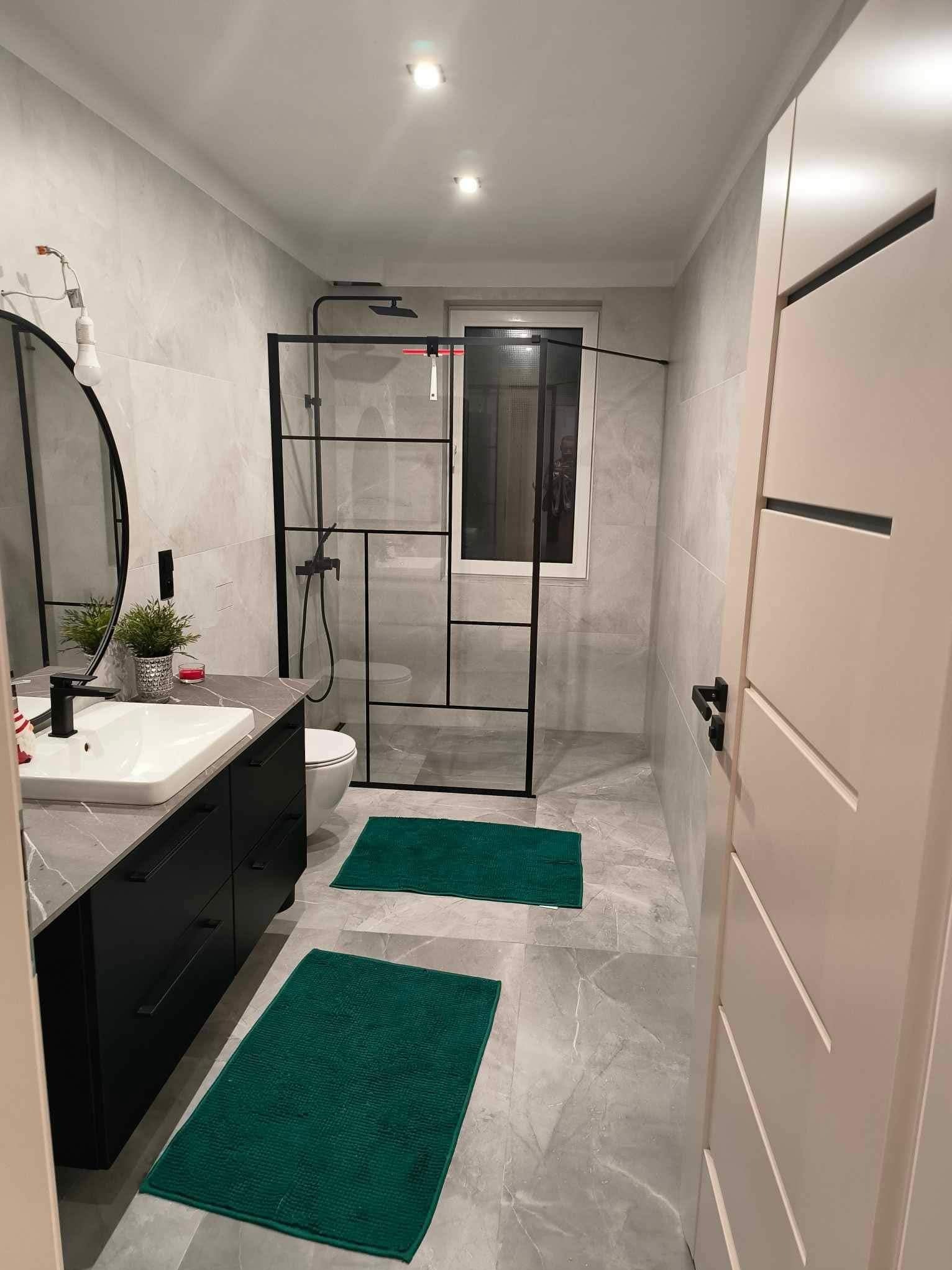 Nowoczesna łazienka z szarymi płytkami, kabiną prysznicową typu walk-in z czarnymi profilami, czarną szafką pod umywalką i zielonymi dywanikami.