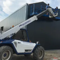 Dwóch monterów na podnośniku koszowym instaluje ciemnoszare płyty warstwowe na ścianie powstającej hali stalowej, widoczny fragment logo firmy na maszynie.