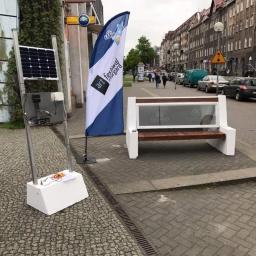 Promad fotowoltaika Stargard - Uliczna instalacja: panel fotowoltaiczny na metalowym stelażu z widocznym okablowaniem, obok ławka z panelem reklamowym na tle miejskiej ulicy ze starą zabudową.