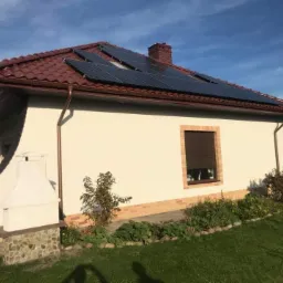 Instalacja paneli słonecznych na dachu domu jednorodzinnego z brązową dachówką i kremową elewacją, widok z poziomu ogrodu, w tle błękitne niebo.