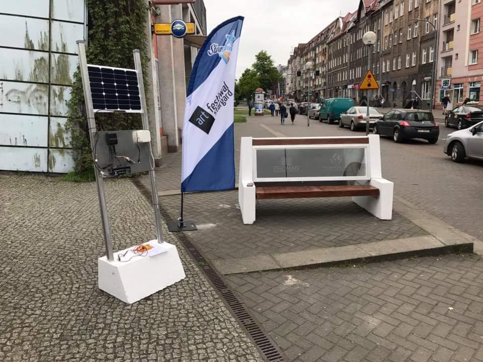 Uliczna instalacja: panel fotowoltaiczny na metalowym stelażu z widocznym okablowaniem, obok ławka z panelem reklamowym na tle miejskiej ulicy ze starą zabudową.