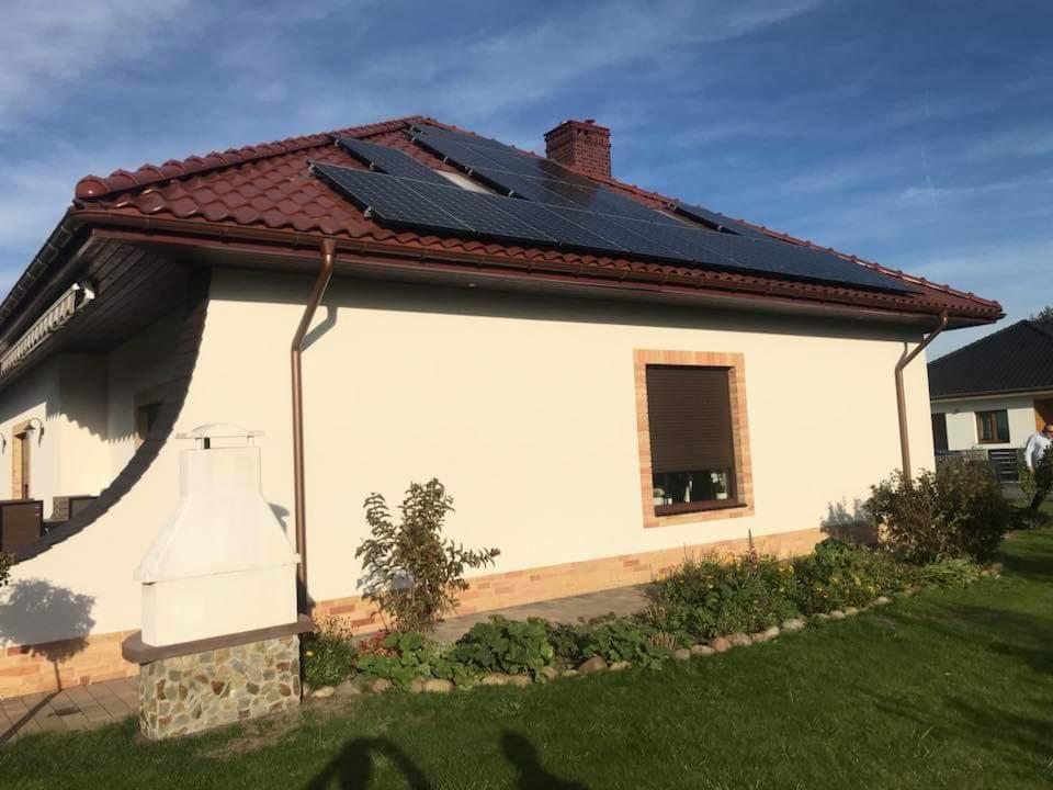 Instalacja paneli słonecznych na dachu domu jednorodzinnego z brązową dachówką i kremową elewacją, widok z poziomu ogrodu, w tle błękitne niebo.