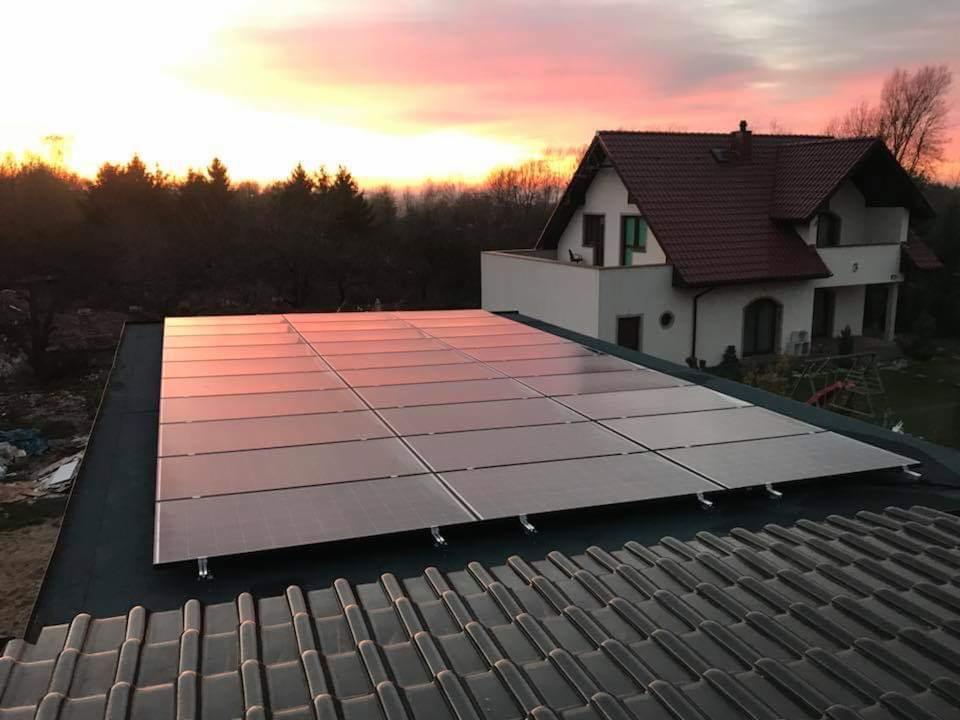 Panel słoneczny na dachu domu jednorodzinnego, odbijający promienie zachodzącego słońca, z widoczną dachówką w pierwszym planie i drzewami w tle.