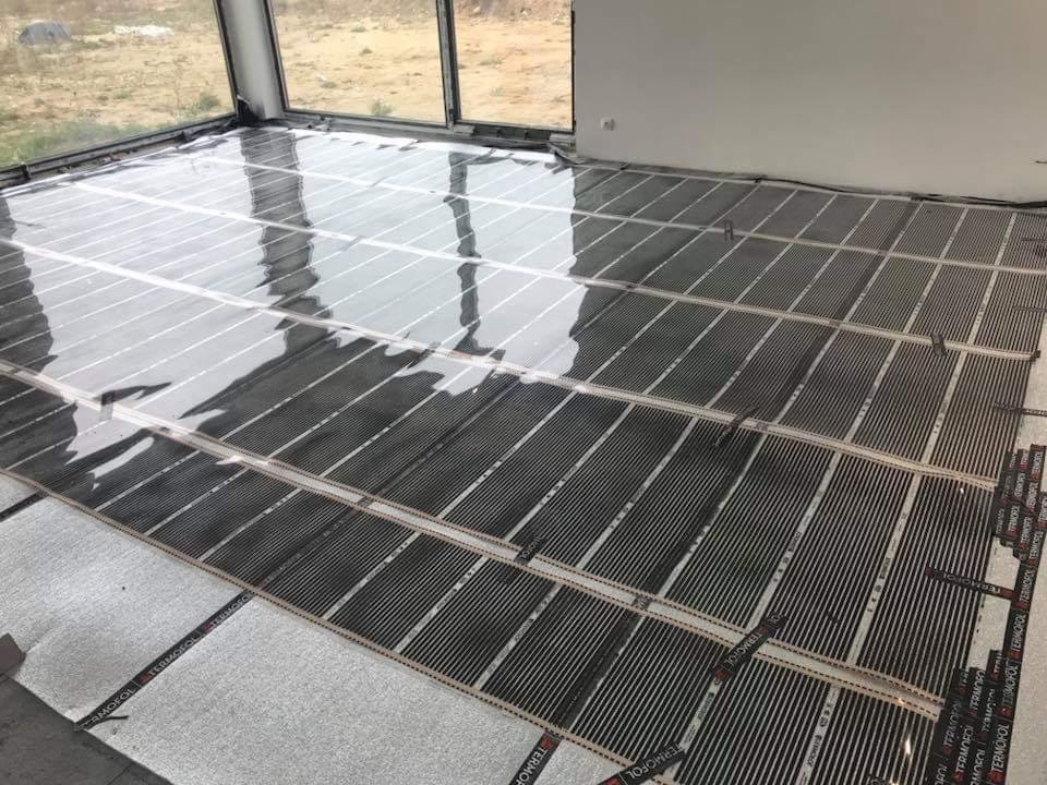 Rozłożona mata grzewcza Termofol na podkładzie izolacyjnym w pustym, jasnym pomieszczeniu z dużymi oknami, przygotowana do montażu ogrzewania podłogowego.