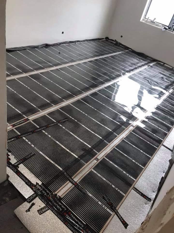 Rozłożona folia grzewcza Termofol na podłodze, przygotowana do montażu, widoczne srebrne podłoże i czarne pasy grzewcze, białe ściany w tle.