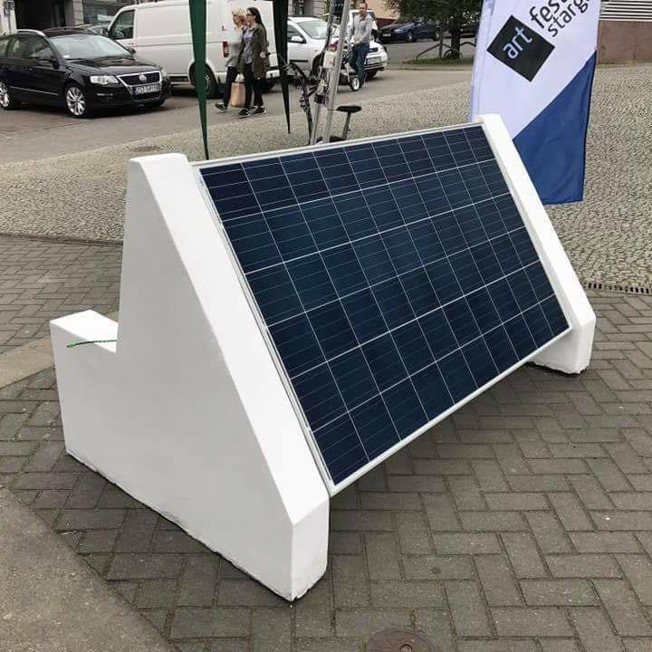 Przenośny panel słoneczny z białym betonowym stojakiem ustawiony na brukowanej ulicy, flaga festiwalowa w tle.