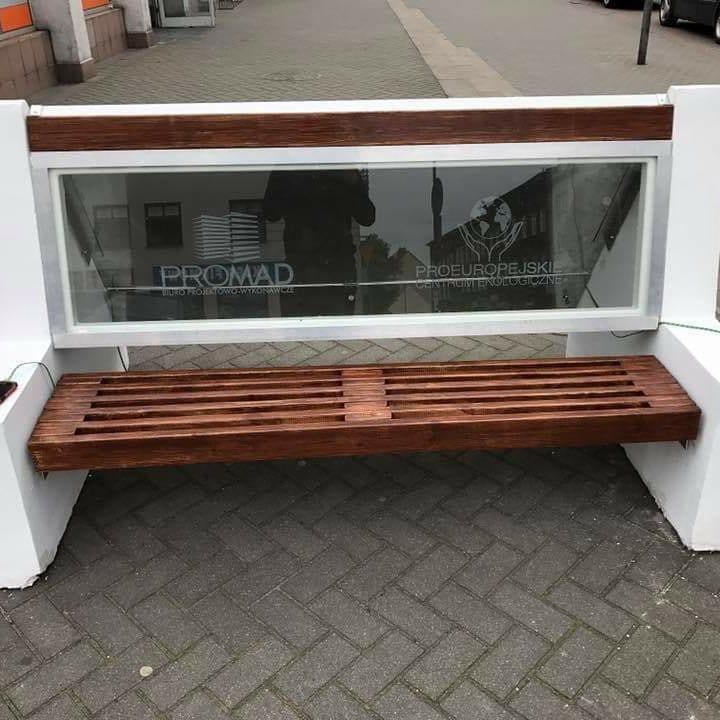 Ławka z drewnianym siedziskiem i przeszklonym oparciem, na którym widnieją logotypy firm Promad i Proeuropejski Centrum Ekologiczne, ustawiona na chodniku z kostki.