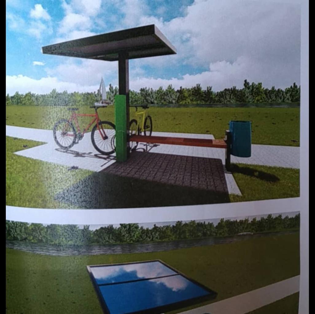 Wizualizacja projektu stacji ładowania rowerów z panelem fotowoltaicznym na zadaszeniu, obok stoją rowery: czerwony i żółty, w tle zielony krajobraz.