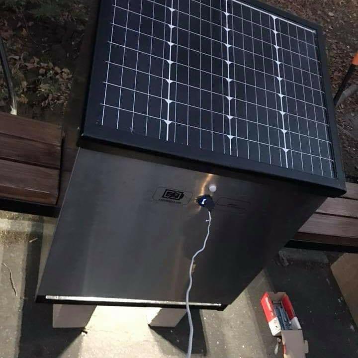 Zewnętrzna ławka solarna z panelem fotowoltaicznym, gniazdem USB i oświetleniem LED, umieszczona na betonowej nawierzchni obok drewnianego siedziska.