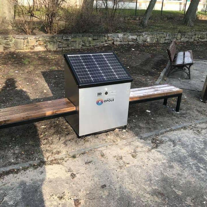 Ławka parkowa z panelem słonecznym na metalowej obudowie, logo Opole, częściowo pokryta śniegiem, na tle drzew i kamiennego murku.