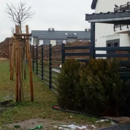 Nowoczesne, poziome ogrodzenie panelowe w kolorze antracytowym, widok z zewnątrz posesji, na tle budowy i ciężarówki wywożącej ziemię.