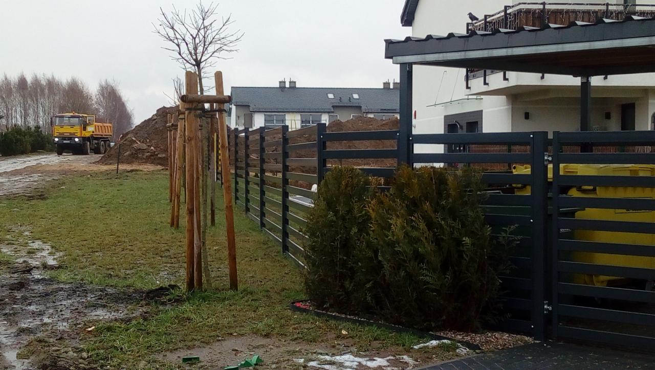 Nowoczesne, poziome ogrodzenie panelowe w kolorze antracytowym, widok z zewnątrz posesji, na tle budowy i ciężarówki wywożącej ziemię.