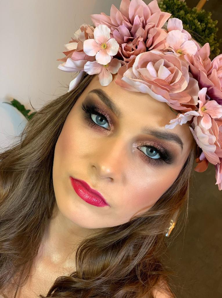 Portret kobiety z intensywnym, wieczorowym eye-art z użyciem brązów i błysku, podkreślonymi rzęsami, różowymi ustami i delikatnym różem na policzkach, a także wiankiem z różowych kwiatów na głowie.