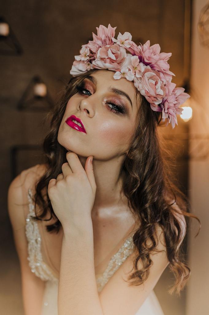 Portret kobiety z błyszczącym cieniem do powiek w odcieniach różu i fuksjową szminką, noszącej wianek z różowych kwiatów i sukienkę z koronkowymi zdobieniami.