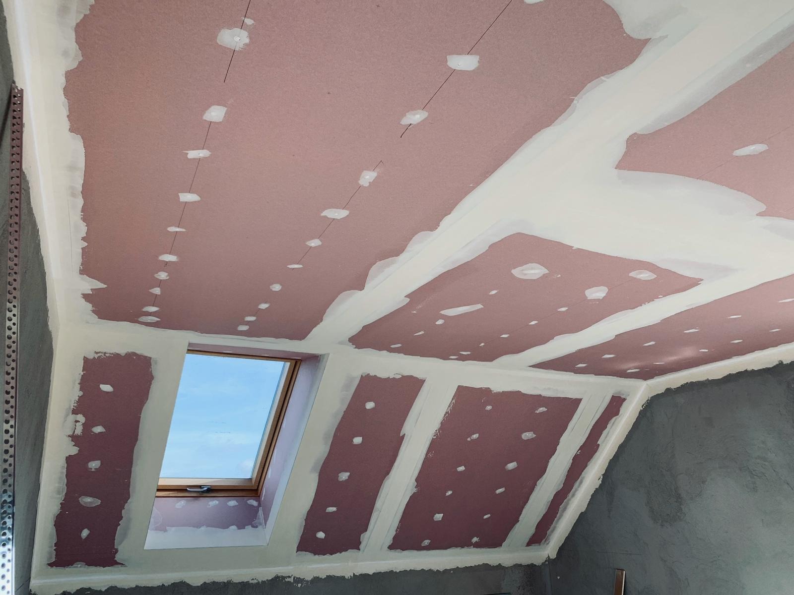 Montaż płyt kartonowo-gipsowych na poddaszu z oknem dachowym, widoczne szpachlowane łączenia i punkty mocowania, surowe ściany w tle.