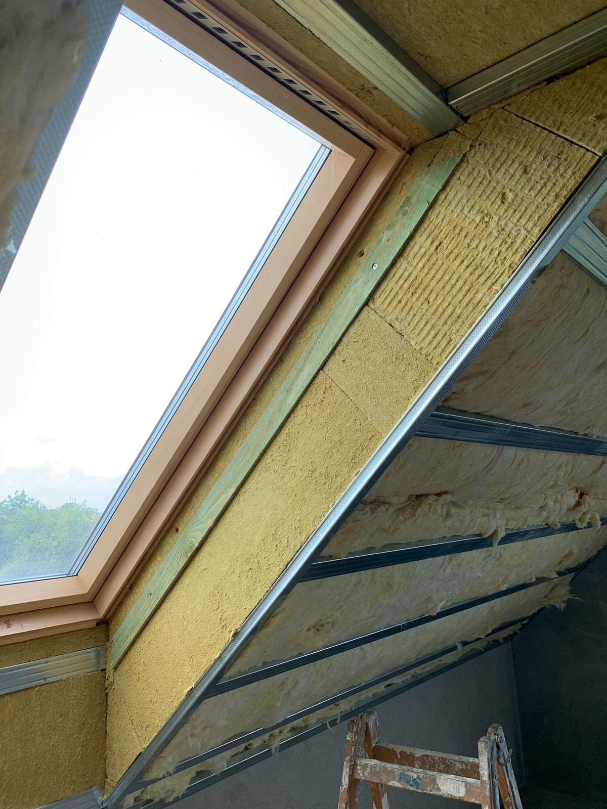 Montaż okna dachowego z izolacją wełną mineralną, widoczne metalowe profile i drewniane elementy konstrukcyjne, drabina oparta o ścianę.