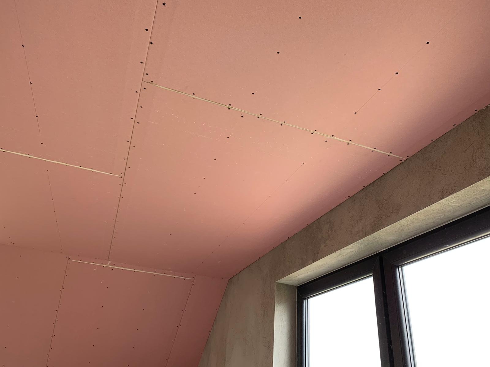 Różowa okładzina sufitowa zamocowana do stelaża, widoczne wkręty, surowy tynk na ścianie i rama okienna.