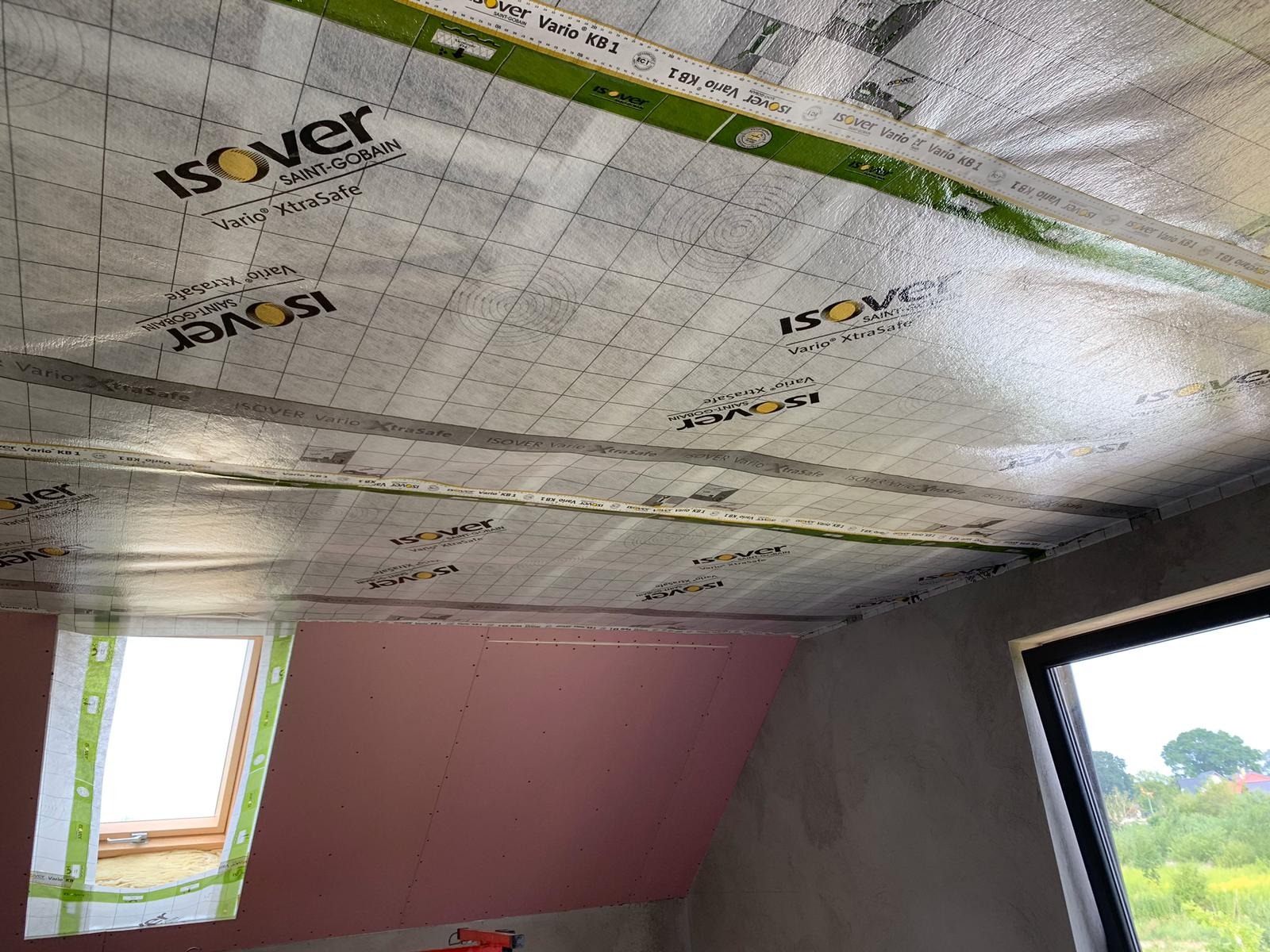 Montaż izolacji folią paroizolacyjną Isover Vario XtraSafe na poddaszu z oknem dachowym, widoczne fragmenty ścian obłożonych płytami w kolorze różowym.