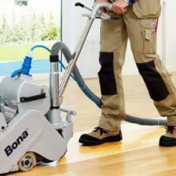 Szlifowanie drewnianej podłogi parkieciarką Bona przez pracownika w beżowych spodniach roboczych z czarnymi wzmocnieniami na kolanach i brązowych butach roboczych.