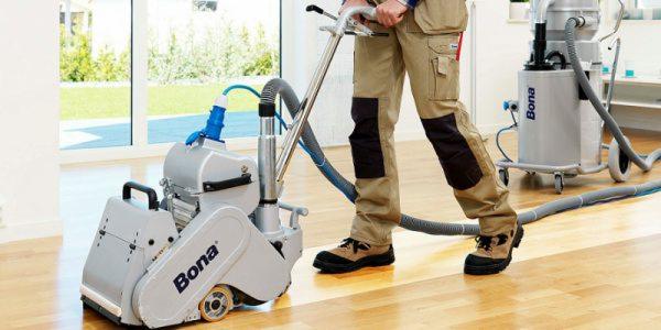 Szlifowanie drewnianej podłogi parkieciarką Bona przez pracownika w beżowych spodniach roboczych z czarnymi wzmocnieniami na kolanach i brązowych butach roboczych.