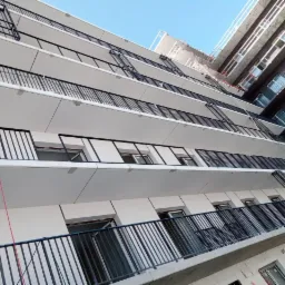 Montaż czarnych, metalowych balustrad balkonowych w nowym bloku mieszkalnym, widok z dołu na elewację z pracownikiem na linie zabezpieczającej.