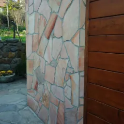 Fragment elewacji budynku z okładziną z nieregularnych, różowo-beżowych kamieni, przylegający do drewnianej ściany z poziomych desek, z widoczną kamienną ścieżką i roślinnością w tle.