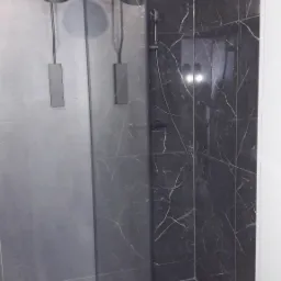 Nowoczesna kabina prysznicowa z czarnymi płytkami imitującymi marmur i czarną armaturą, oświetlona industrialną lampą ścienną