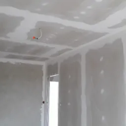 Wnętrze w trakcie prac wykończeniowych: surowe ściany i sufit z płyt kartonowo-gipsowych, widoczne łączenia i punkt oświetleniowy z wystającymi przewodami, przygotowane do dalszych etapów wykończenia.