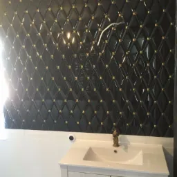 Błyszczące, czarne płytki ścienne z wypukłym wzorem rombów i złotymi detalami, nad białą umywalką z baterią w stylu retro, widoczne przewody elektryczne.