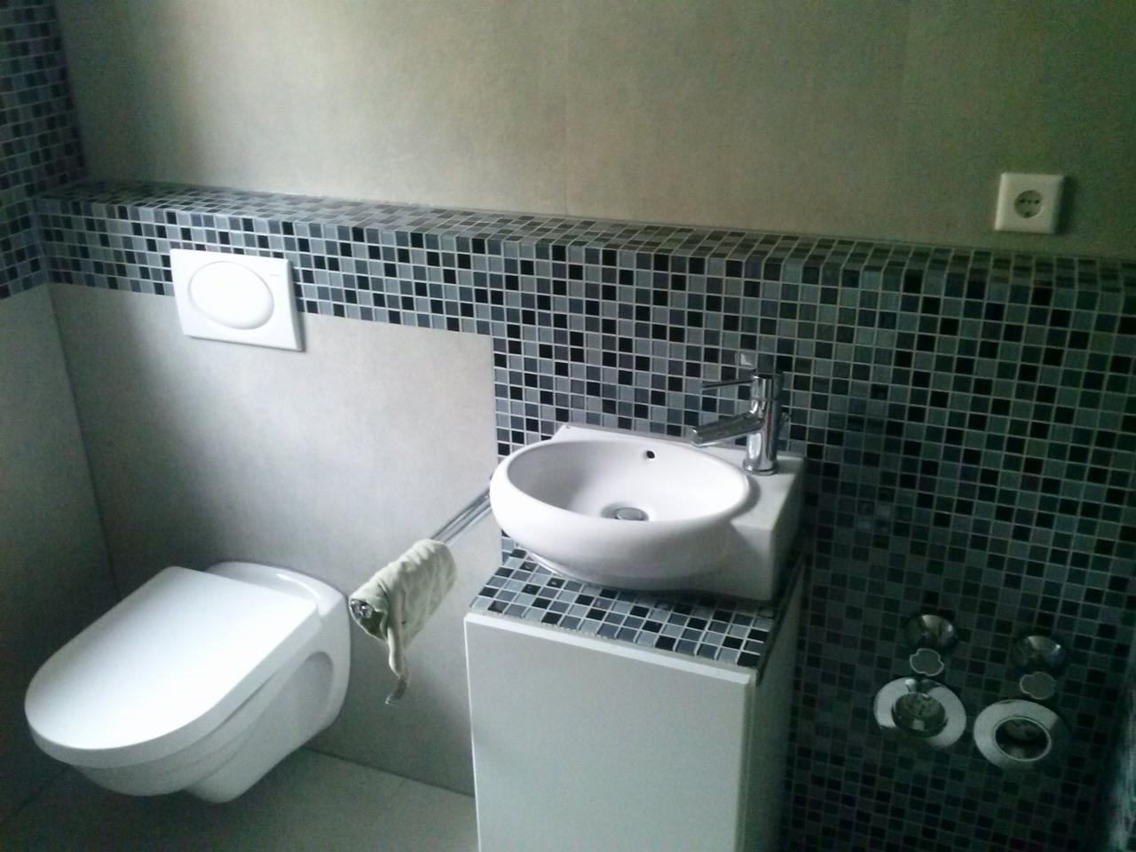 Nowoczesna łazienka z wiszącą toaletą, umywalką nablatową na szafce i mozaiką w odcieniach szarości na ścianach.