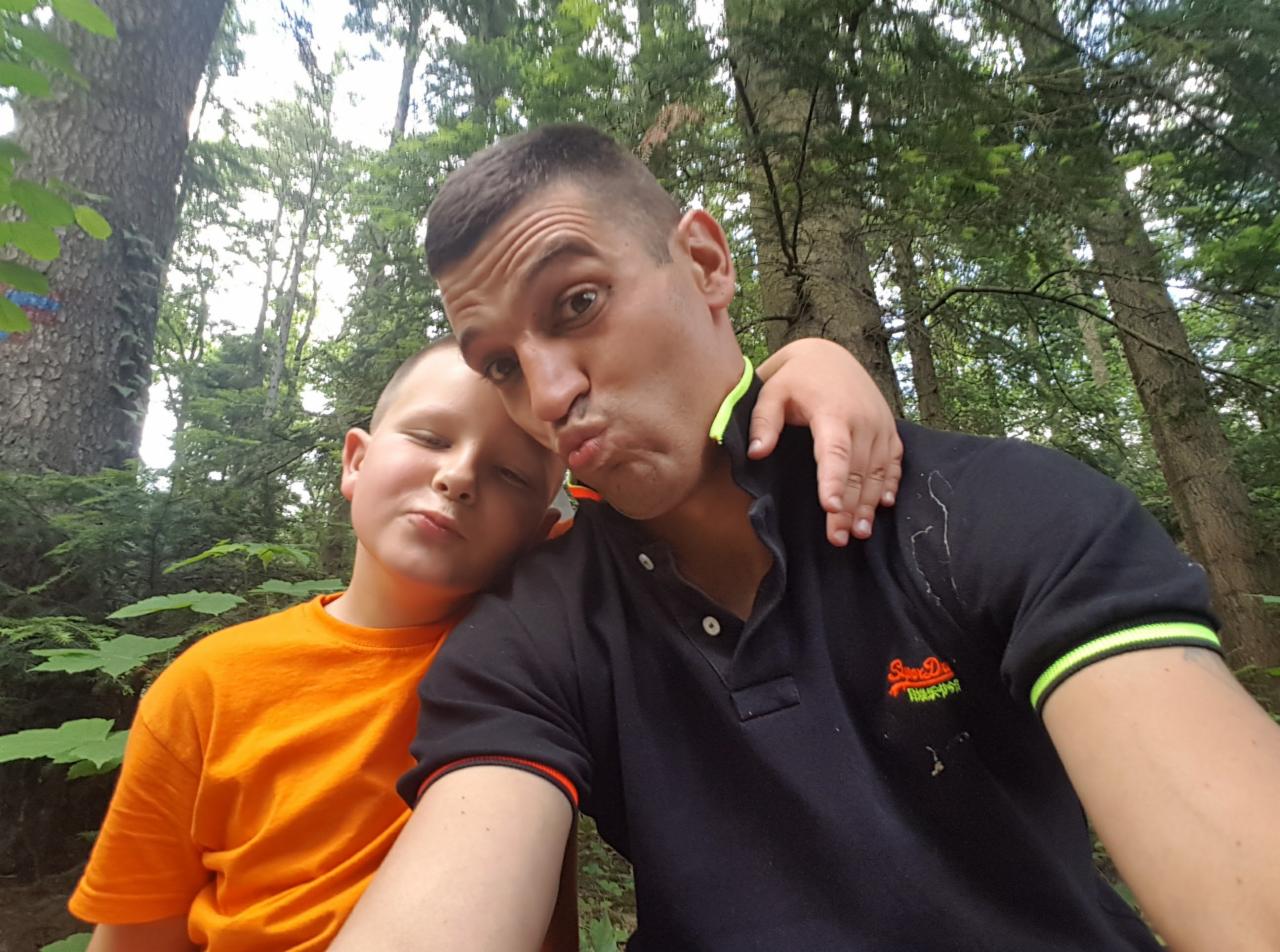 Selfie ojca i syna w lesie. Mężczyzna w czarnej koszulce polo robi zabawną minę, obejmując syna w pomarańczowej koszulce. W tle drzewa i zielona roślinność.