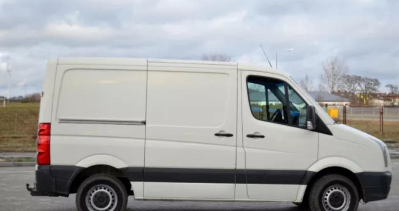 Biały samochód dostawczy typu furgon, widok z boku, na tle pochmurnego nieba i zabudowań.