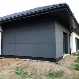 Nowoczesna elewacja budynku jednorodzinnego z szarymi panelami elewacyjnymi, widoczny fragment ocieplenia na sąsiedniej ścianie, teren wokół budynku w trakcie prac wykończeniowych.