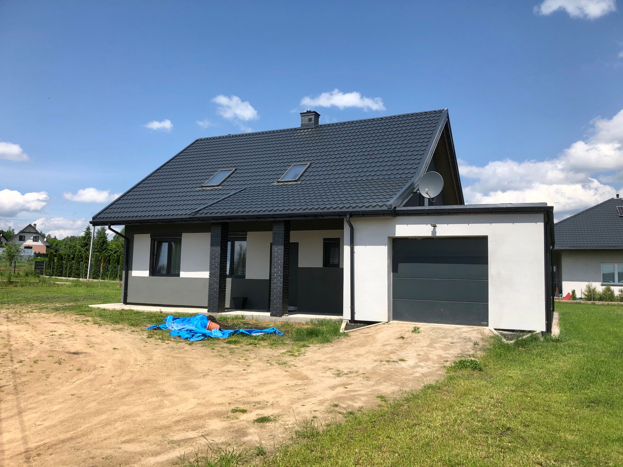 Nowoczesny dom jednorodzinny z szarą elewacją, grafitowym dachem i garażem, widoczna plandeka na podjeździe.