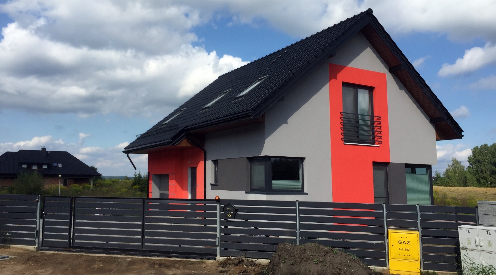 Dwukondygnacyjny dom z szarą elewacją z czerwonymi akcentami wokół okien i czarnym dachem, widoczny zza czarnego, poziomego ogrodzenia.